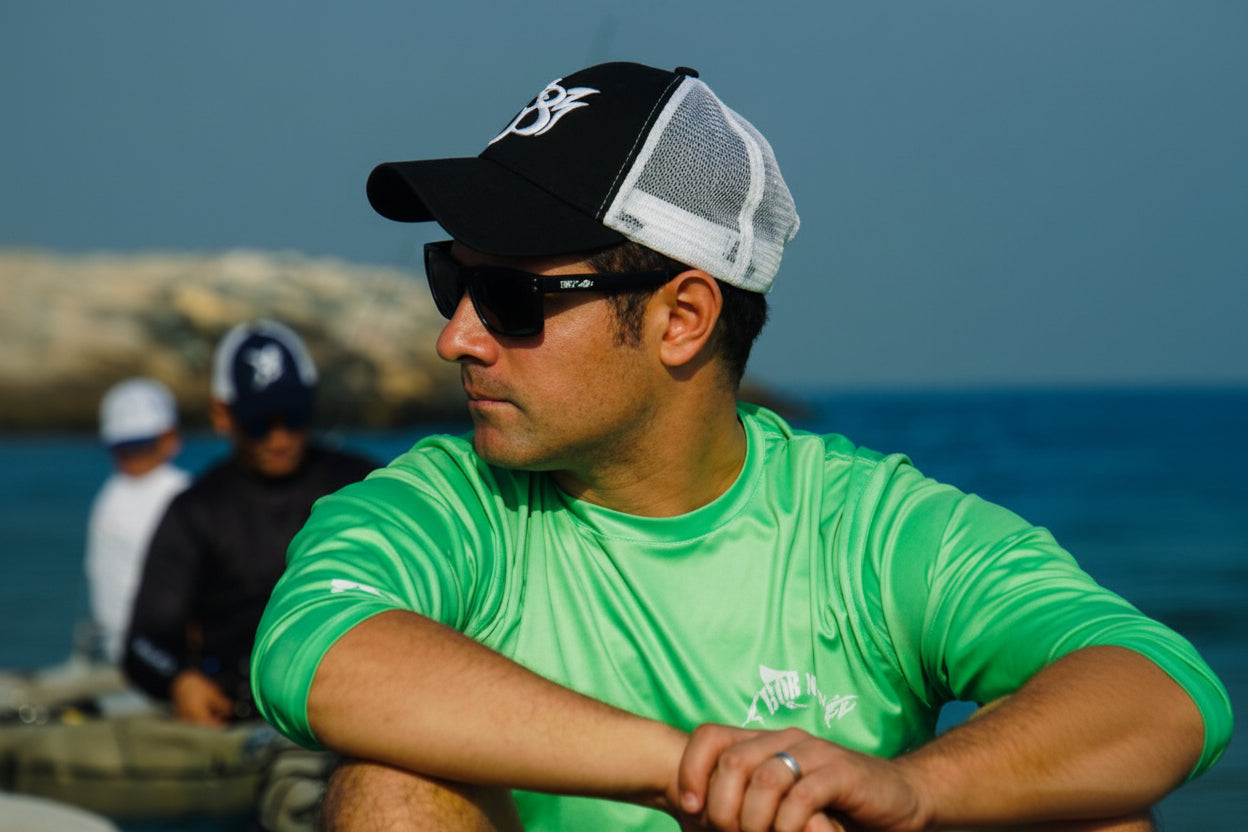 Man wearing a black and white cap and green shirt with sunglasses, sitting outdoors.